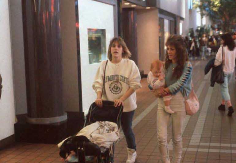 1980s shopping mall hallway with teen mom pushing stroller past storefronts