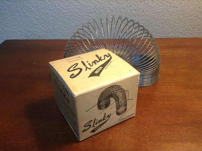 Classic metal Slinky coil standing behind its vintage Slinky box