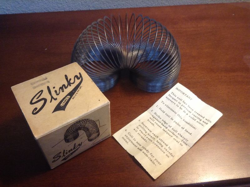 Vintage metal Slinky coil arched beside its original box and instruction sheet